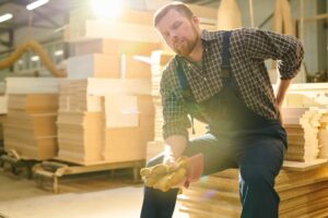 A weary and handsome bearded laborer, eyes closed, sits atop a stack of wooden planks, massaging his lower back to alleviate the pain.